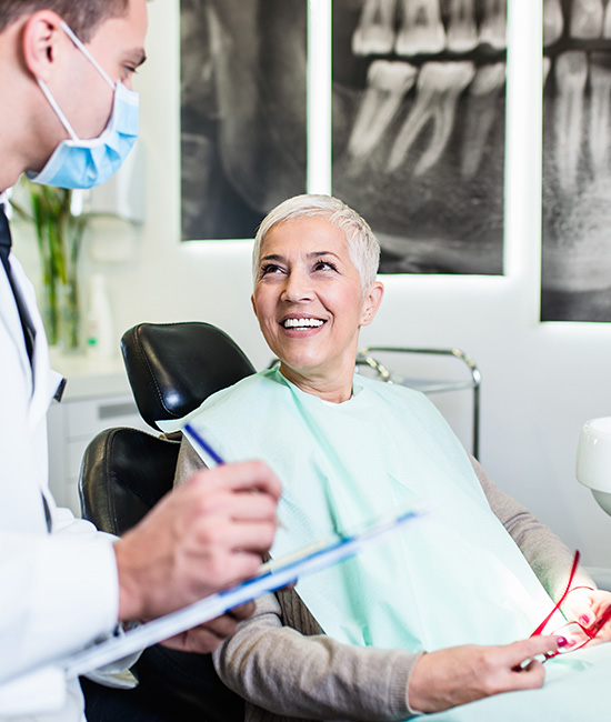 A dental patient is seated in a chair with a smile, receiving attention from a dentist who is standing next to her.